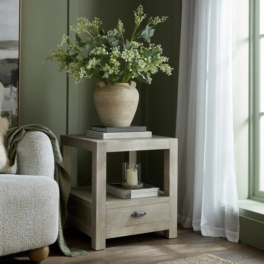 Side table with a floral-patterned lamp and decor items next to a chair in a room with curtains.