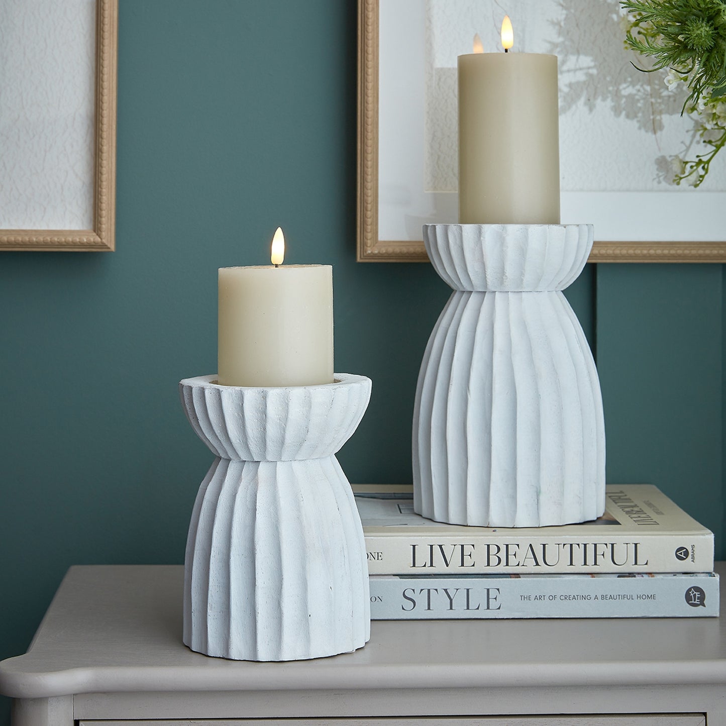 Two white textured candle holders with candles on a surface against a teal wall.
