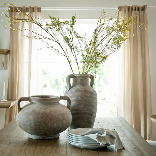 Two large ceramic vases on a wooden table with a window and curtains in the background.