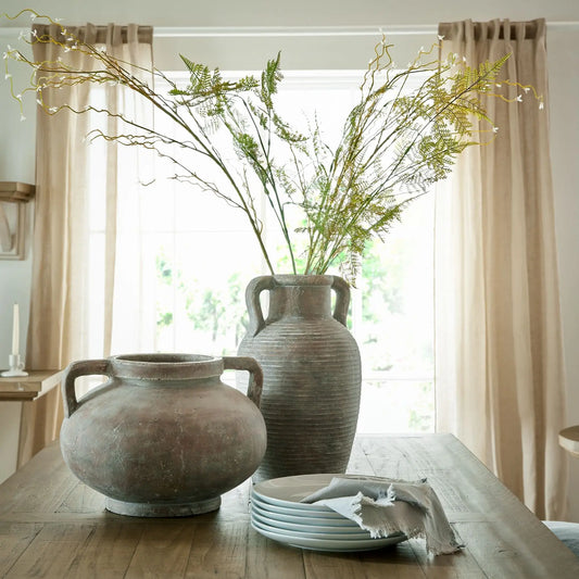 Two large ceramic vases on a wooden table with a window and curtains in the background.