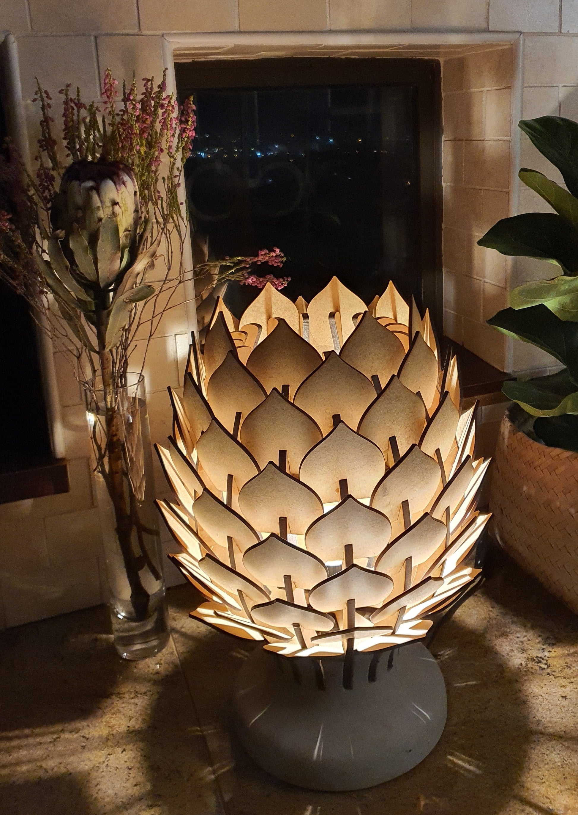 Decorative lamp with a unique design, placed on a surface with a vase of flowers and a plant in the background.