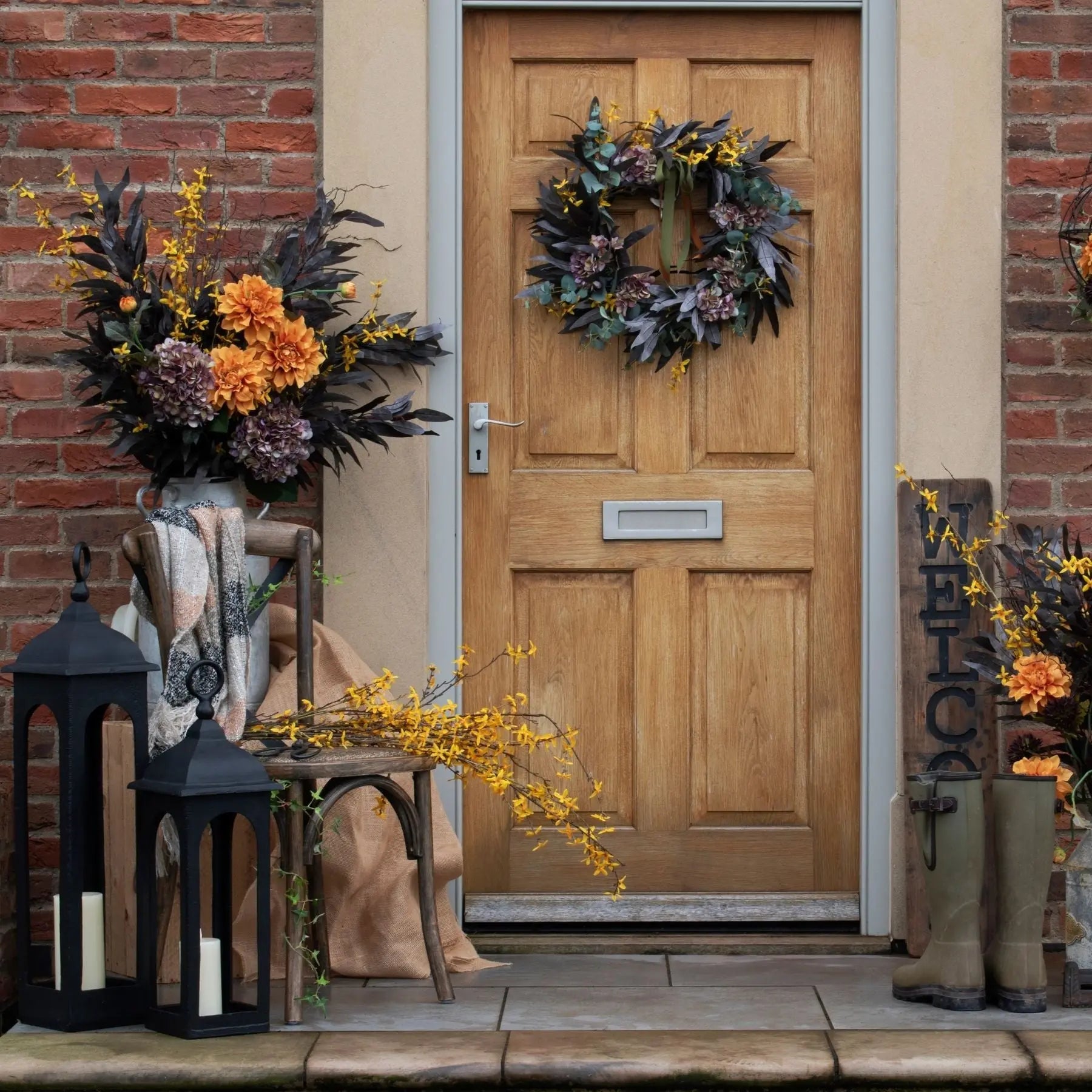 Decorative front door with wreath, lanterns, and flowers on a brick wall.