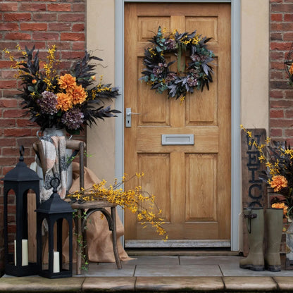 Decorative front door with wreath, lanterns, and flowers on a brick wall.
