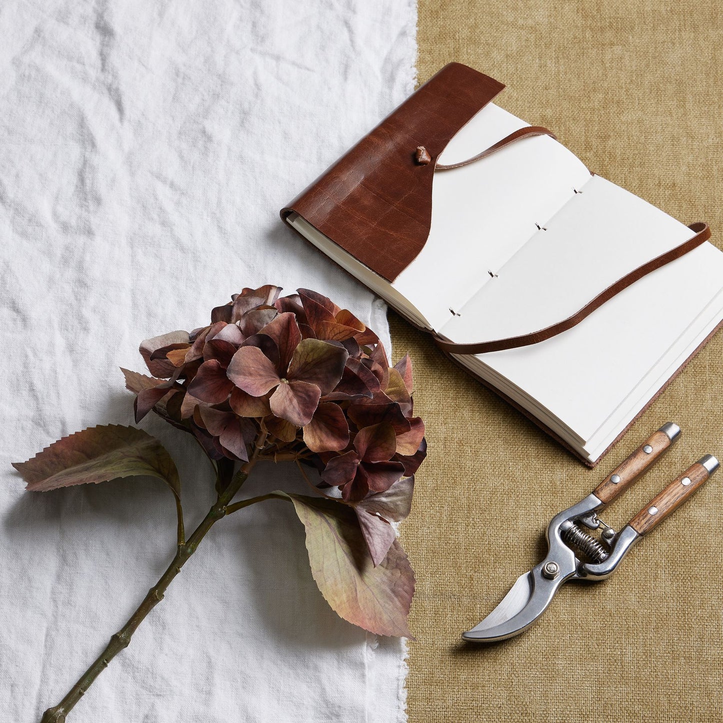 Brown leather notebook with white pages, a dried flower, and garden shears on a textured surface.