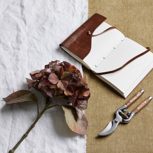 Brown leather notebook with white pages, a dried flower, and garden shears on a textured surface.