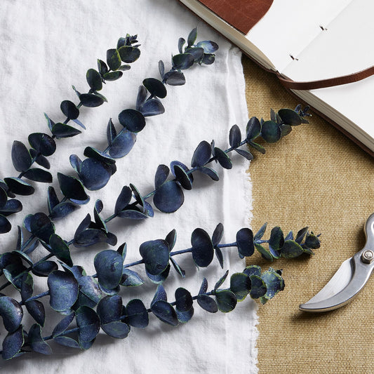Artificial eucalyptus branches on a white surface with a book and scissors in the background.