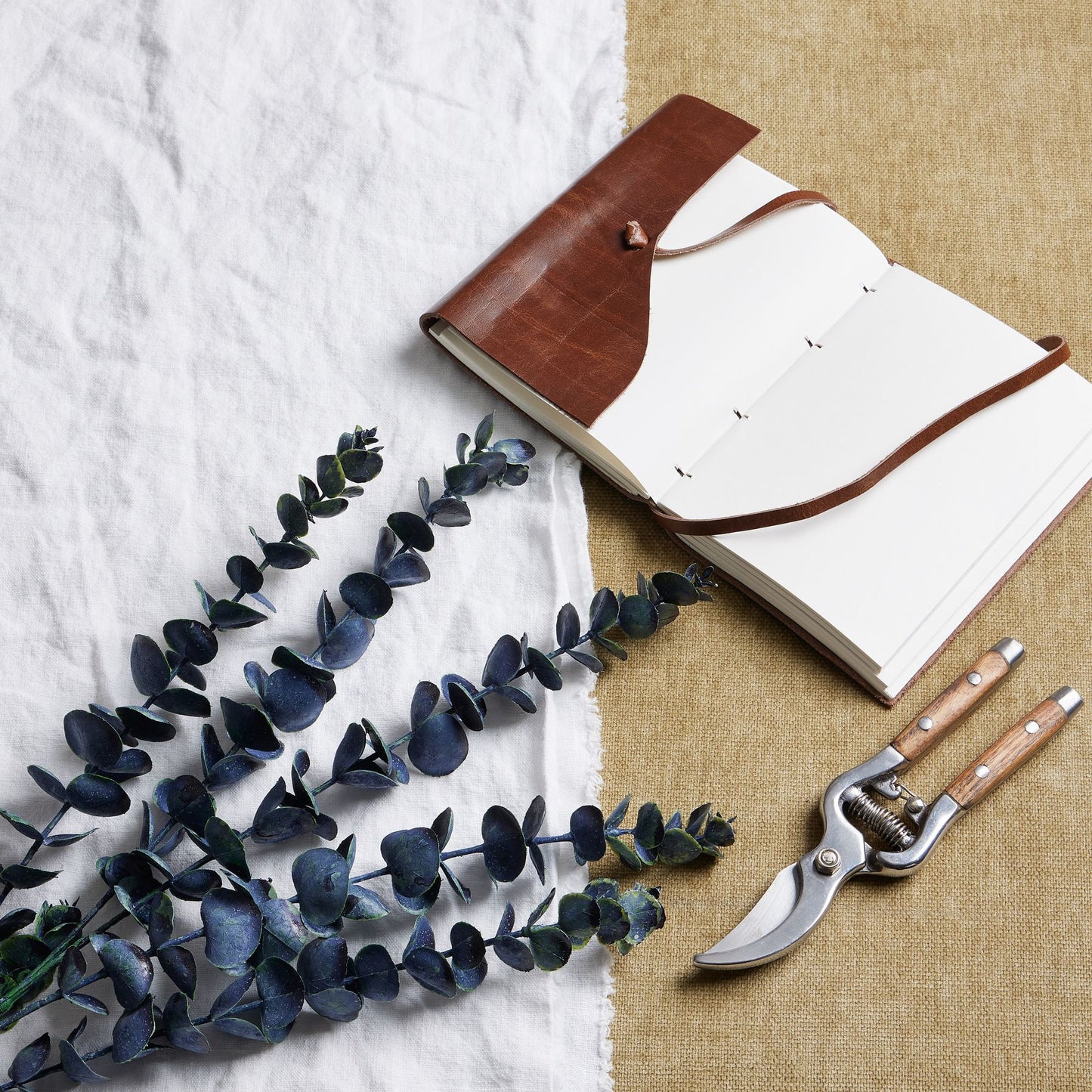 Open notebook with leather binding, eucalyptus branches, and pruning shears on a textured surface.