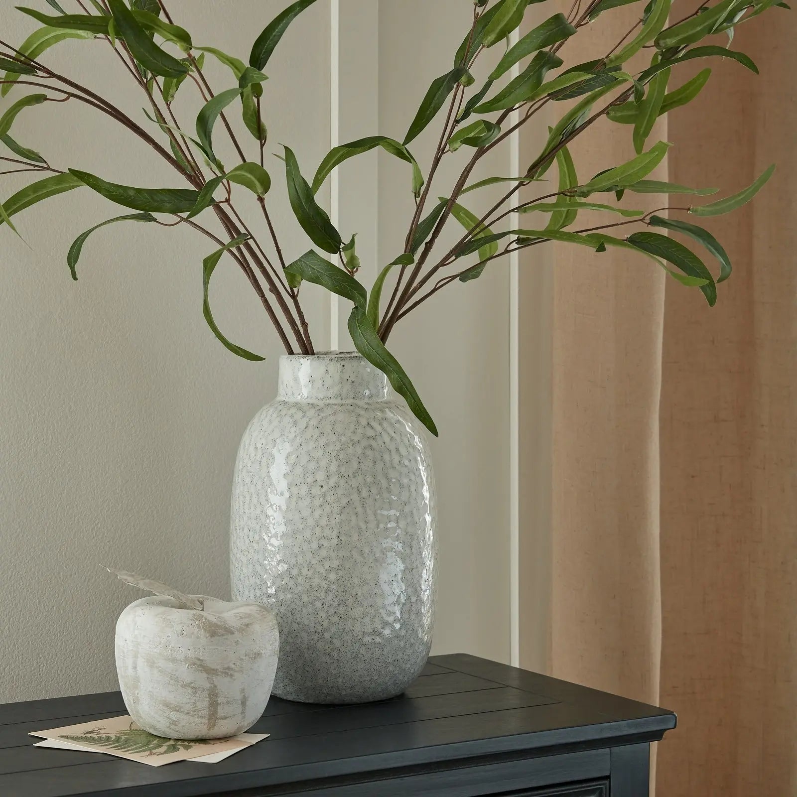 Decorative vase with green branches on a dark surface against a neutral wall.