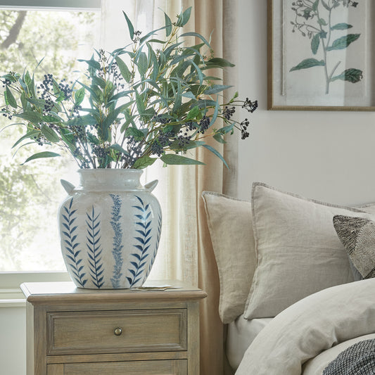 Decorative vase with greenery on a wooden nightstand next to a bed with pillows.