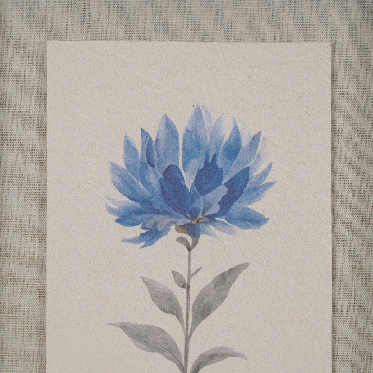 A square art print featuring a watercolor-style blue flower with gray-green leaves on textured off-white paper, displayed against a neutral linen background.