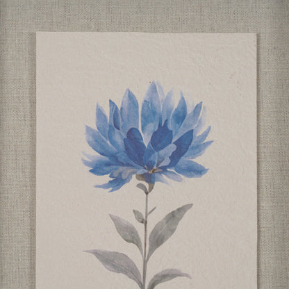 A square art print featuring a watercolor-style blue flower with gray-green leaves on textured off-white paper, displayed against a neutral linen background.