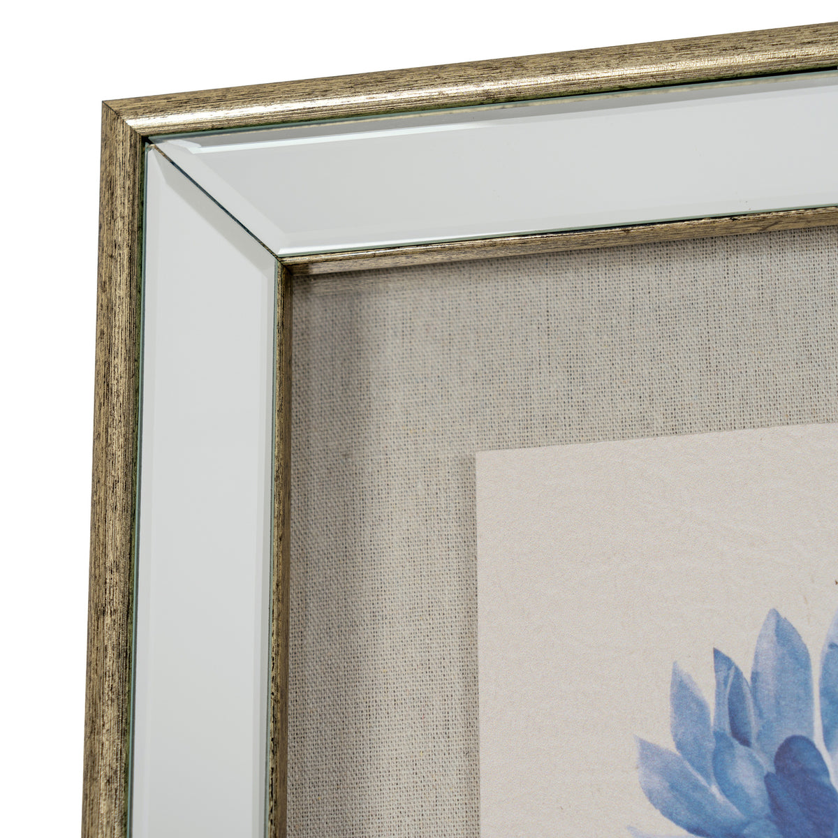 Close-up of a framed wall art corner showing a distressed metallic gold frame, clear glass, textured beige fabric backing, and part of a blue floral print on a light background.