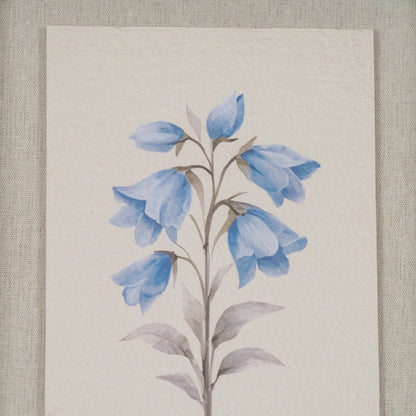 A vertical art print featuring a watercolor painting of blue bell-shaped flowers on a textured off-white paper background, displayed against a neutral beige linen backdrop.