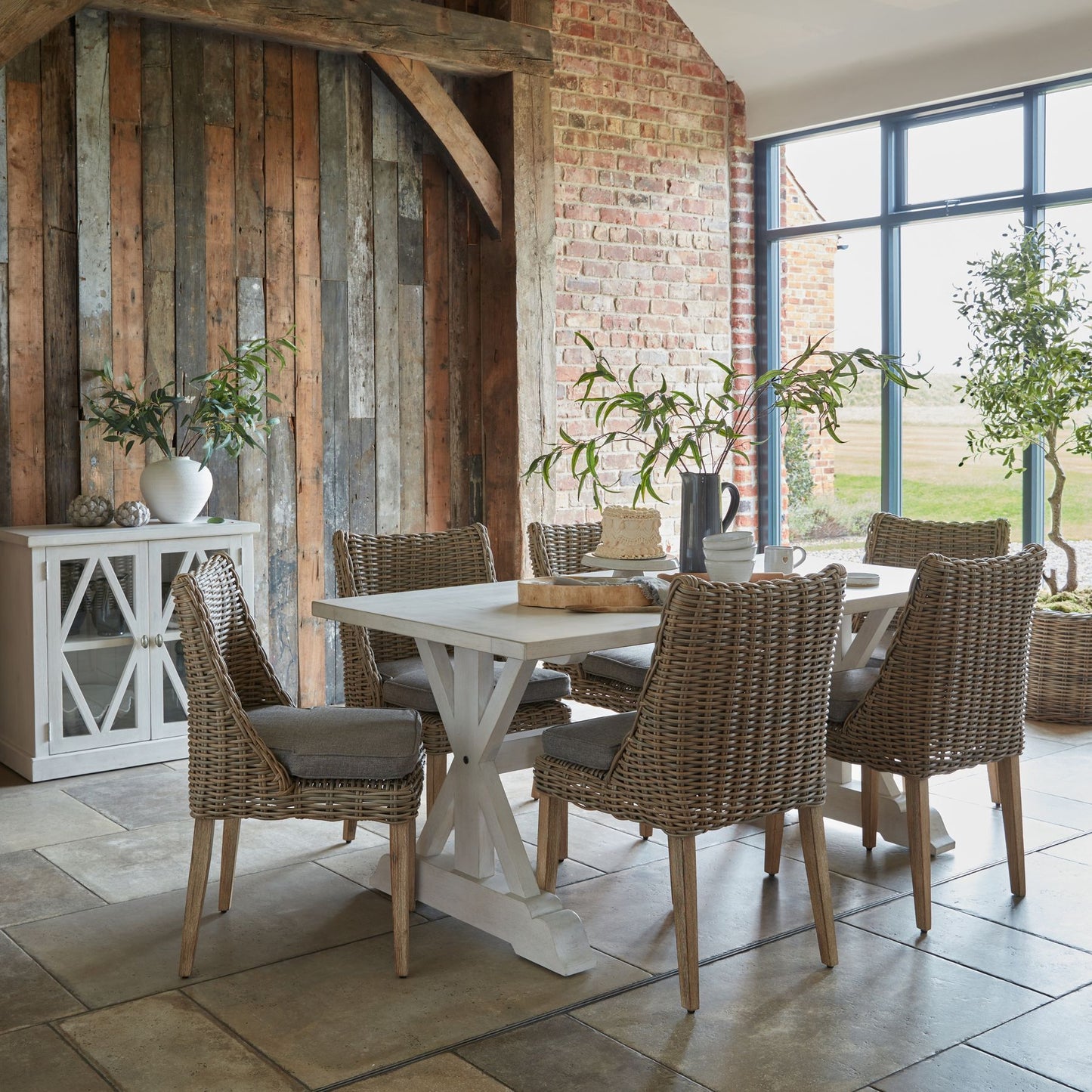 Dining area with a white table and wicker chairs against a rustic brick wall.