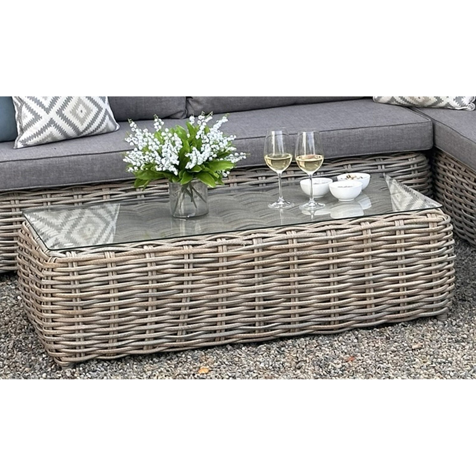 Outdoor patio set with wicker coffee table, cushions, and decorative items on a gravel surface.