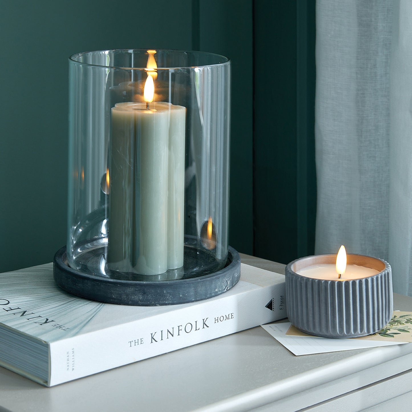 Two candles, one in a glass holder and the other in a ribbed container, on a surface with a book titled 'The Kinfolk Home'.