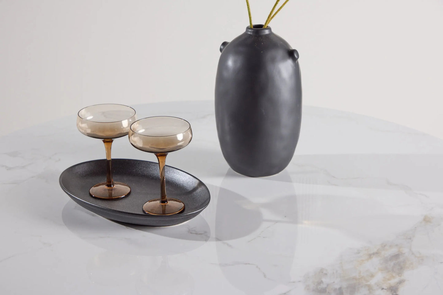 Two glasses with a dark interior on a black plate, accompanied by a dark vase on a light marble surface.