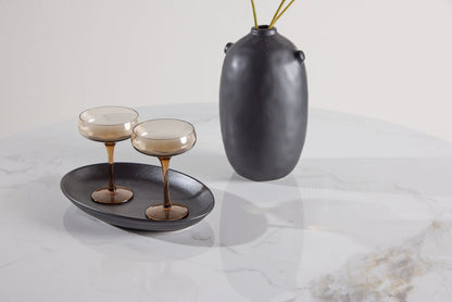 Two glasses with a dark interior on a black plate, accompanied by a dark vase on a light marble surface.