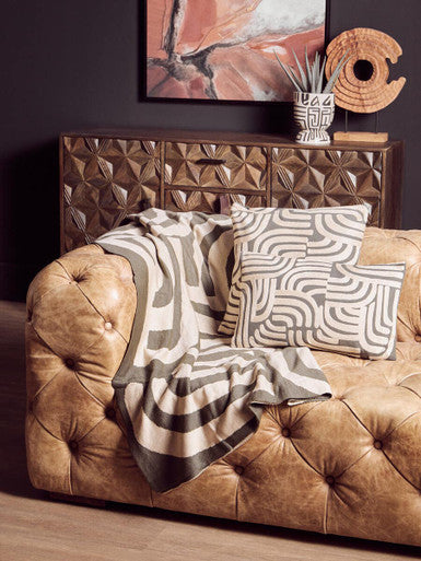 Brown leather sofa with patterned cushions and blanket, against a textured wall with abstract art.