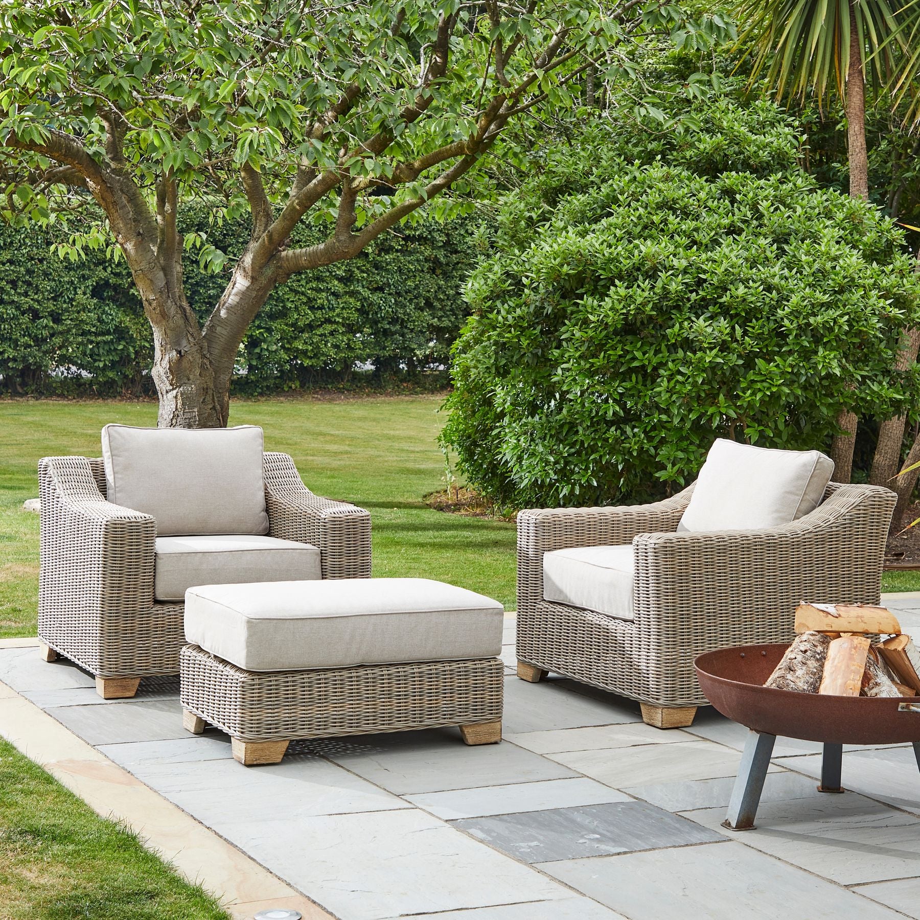 Outdoor patio set with wicker chairs and ottoman on a stone patio, surrounded by greenery.