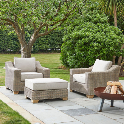 Outdoor patio set with wicker chairs and ottoman on a stone patio, surrounded by greenery.