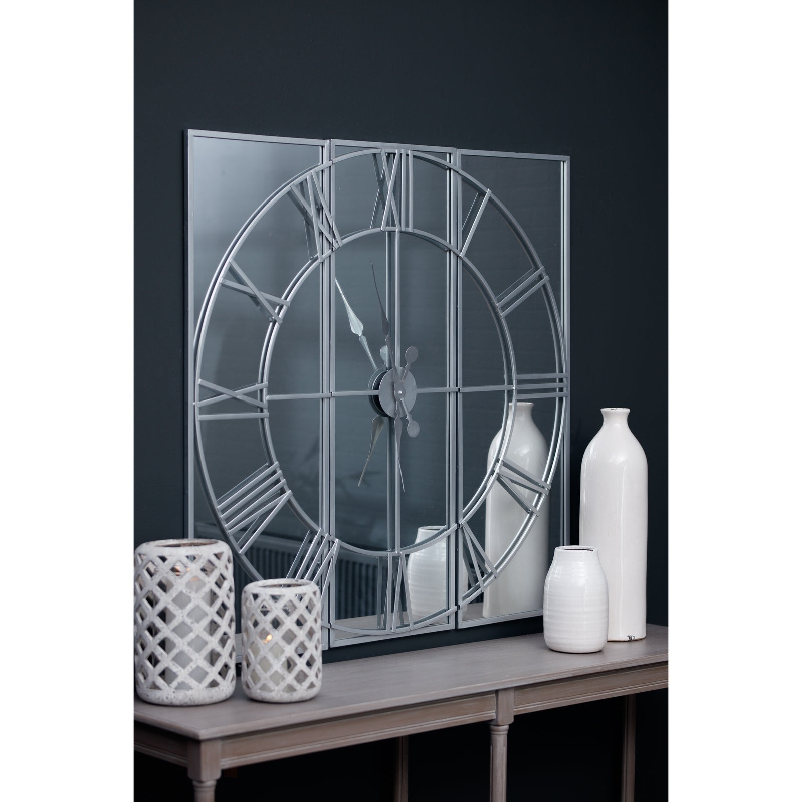 Decorative wall mirror with Roman numeral design on a dark wall with white vases.