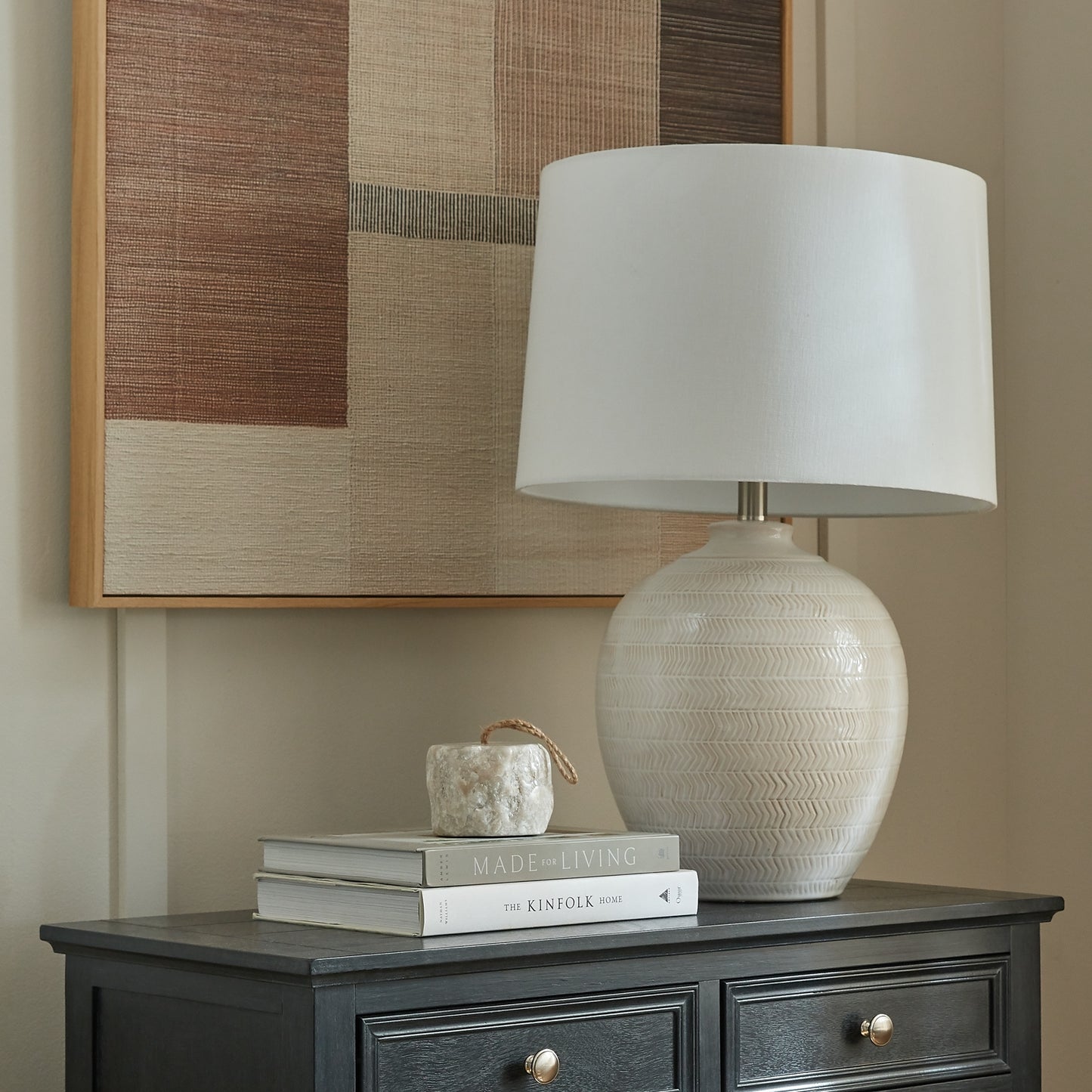 Beige textured lamp on a dark wooden side table with books and a small decorative item, against a neutral wall.
