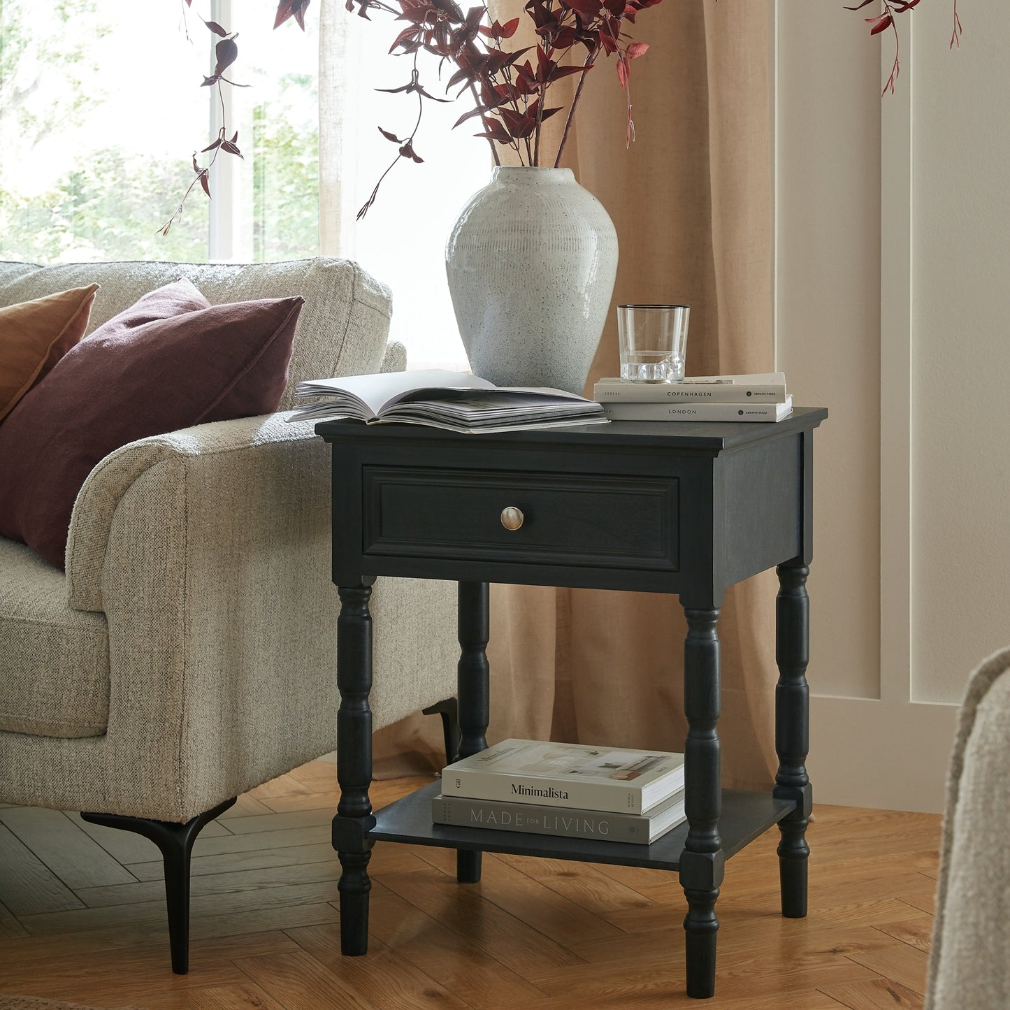 Black side table with books and a vase next to a beige sofa with pillows.