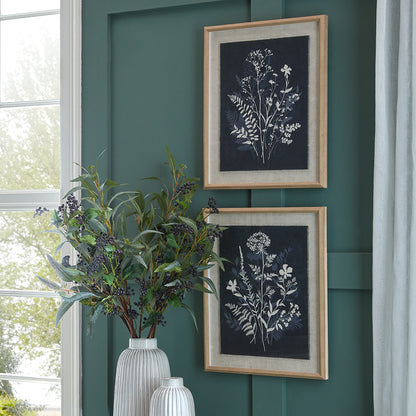 Two framed botanical prints on a green wall with a vase of plants in front.