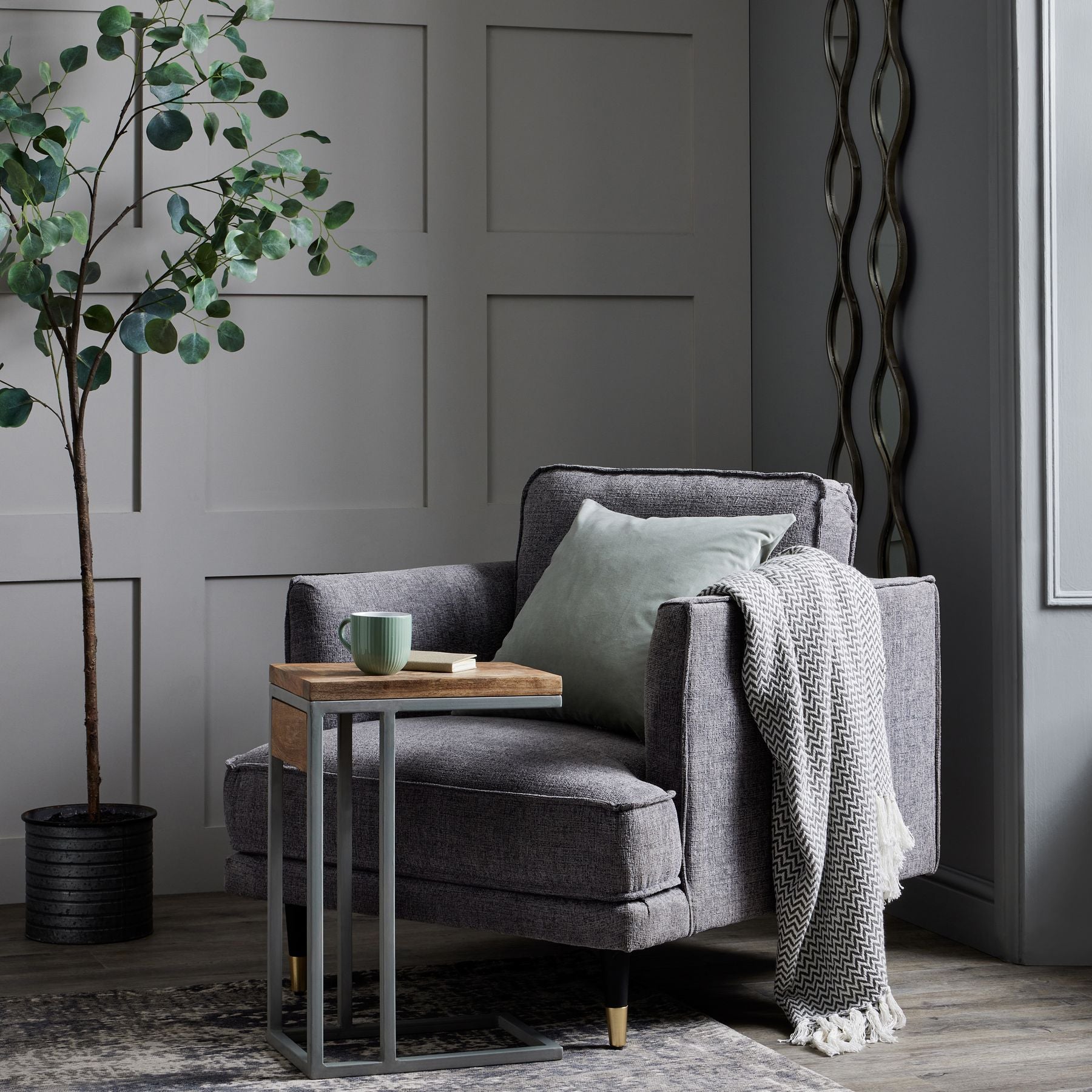 Gray armchair with a wooden side table, green mug, and decorative items in a room with gray walls and a plant.
