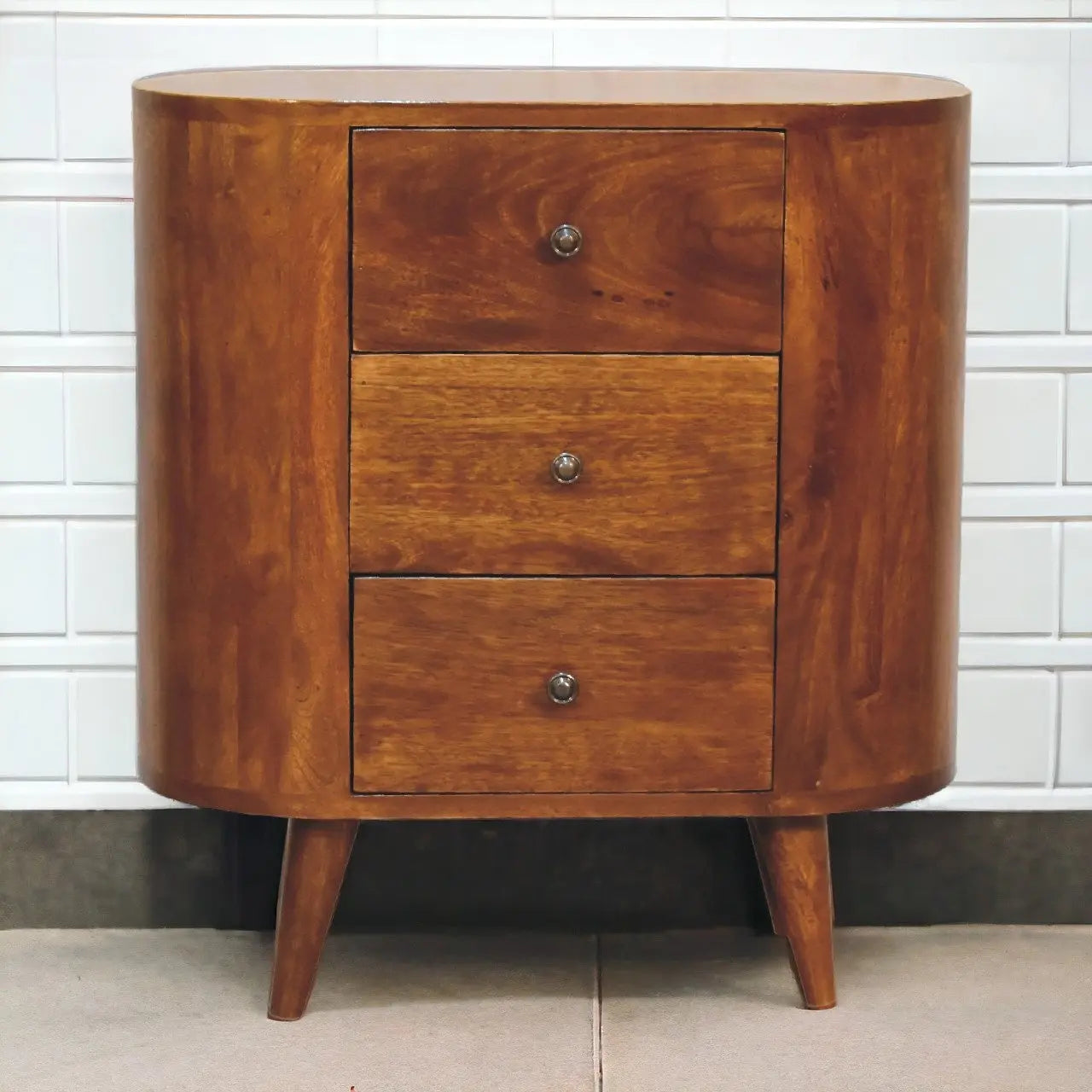 Wooden side table with three drawers against a tiled wall.