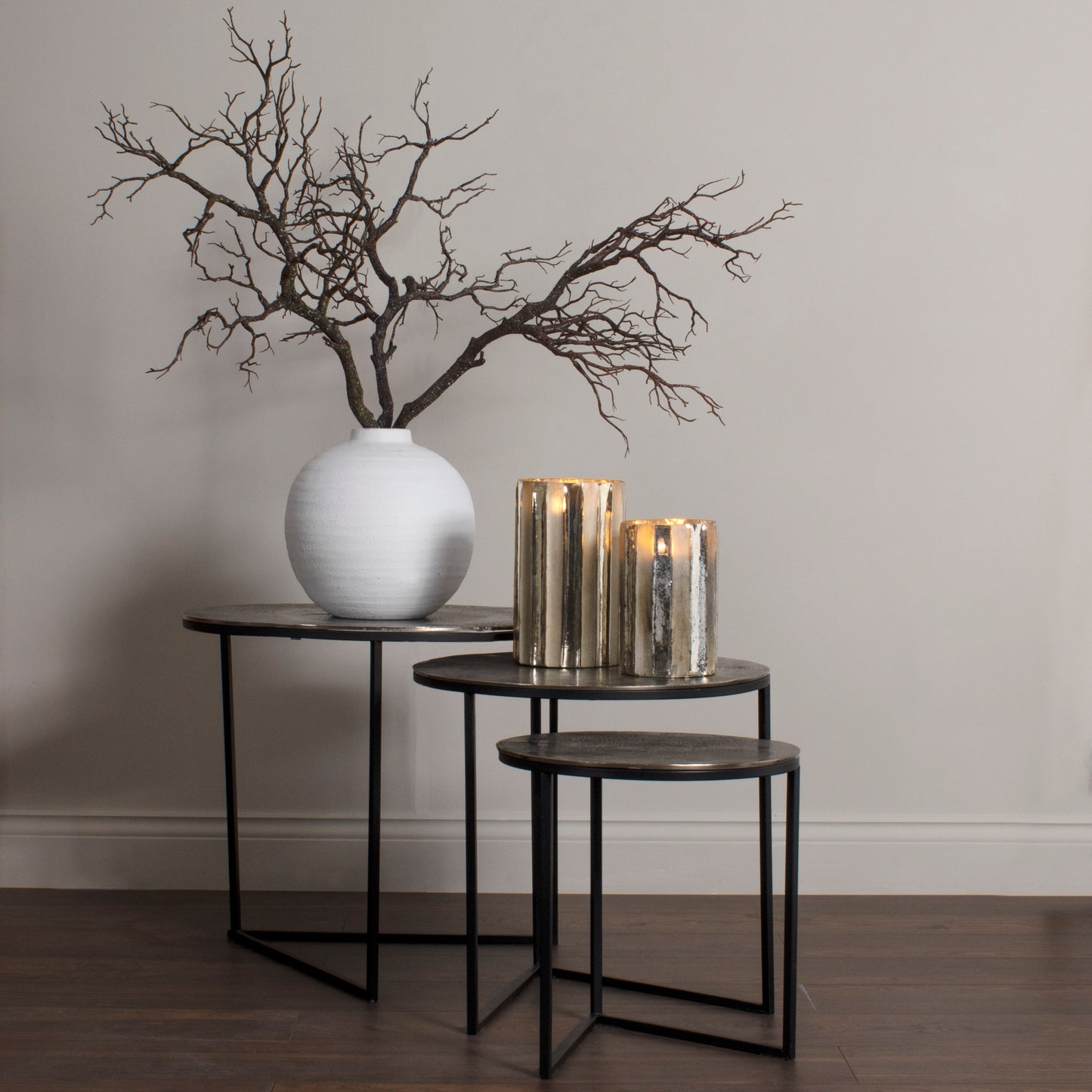 Decorative setup with a white vase, branches, and candles on black tables against a plain wall.