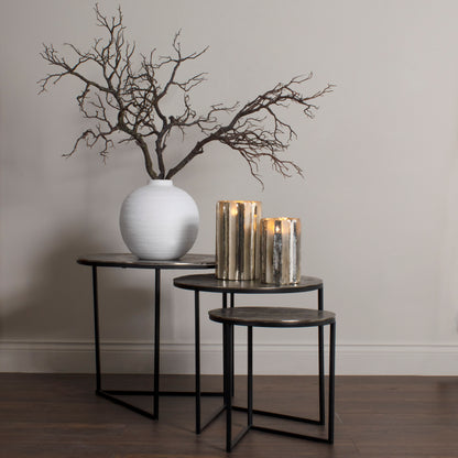 Decorative setup with a white vase, branches, and candles on black tables against a plain wall.