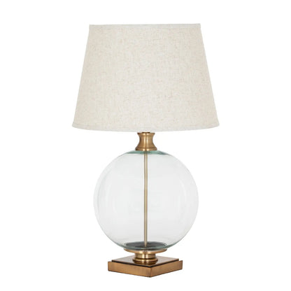 Table lamp with a clear glass base, brass accents, and a beige lampshade on a white background