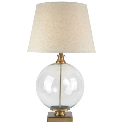 Table lamp with clear glass base, gold accents, and beige fabric shade on a white background