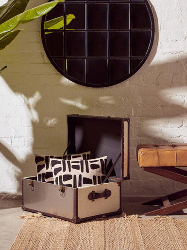 Open suitcase with patterned pillows on a textured rug, against a white brick wall.