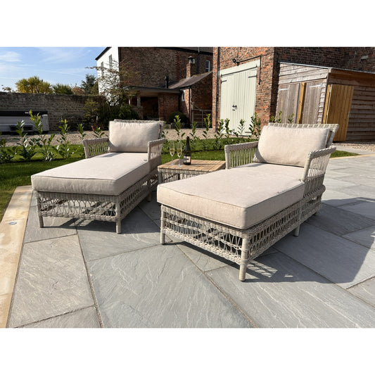 Two wicker outdoor lounge chairs with beige cushions on a stone patio.