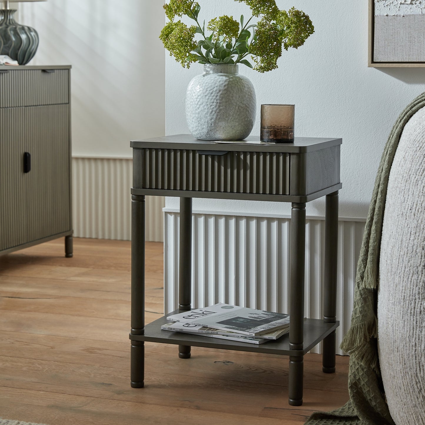 Small dark wood side table with a white textured vase and greenery in a room setting.