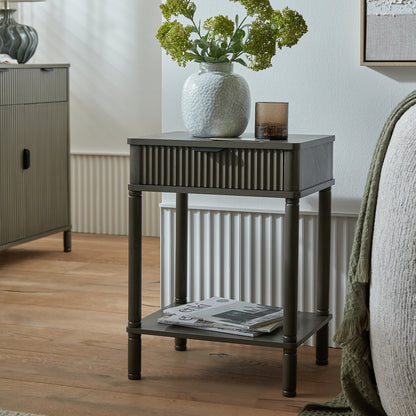Small dark wood side table with a white textured vase and greenery in a room setting.