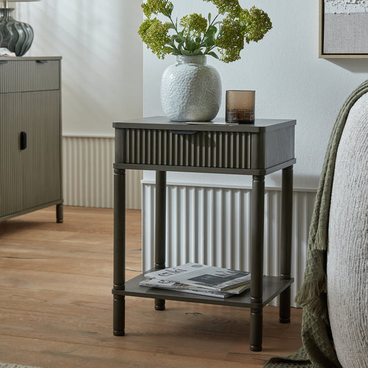 Small dark wood side table with a white textured vase and greenery in a room setting.