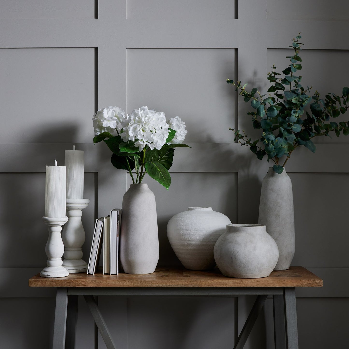 Decorative arrangement of vases, candles, and books on a wooden table against a gray paneled wall.