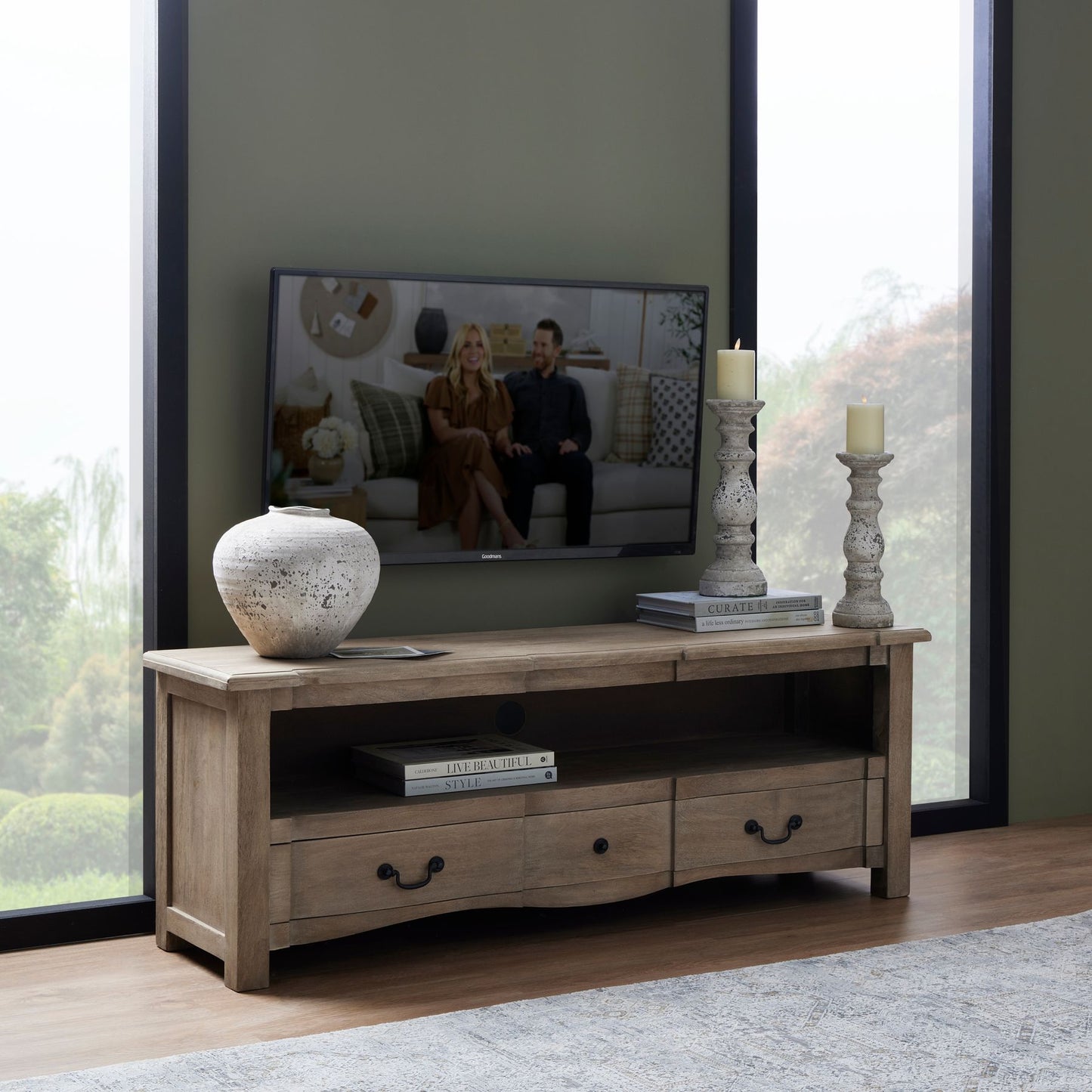 Wooden TV stand with a television displaying two people, surrounded by decorative items in a room with large windows.