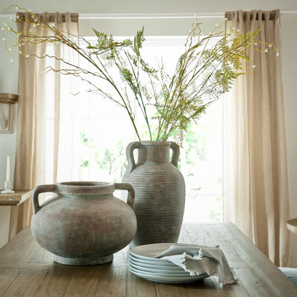 Two large ceramic vases on a wooden table with a window and curtains in the background.
